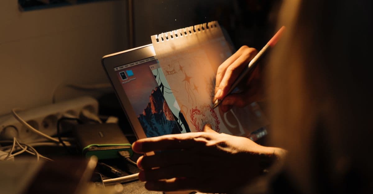 Artist working on a design using a laptop and paper under warm lighting indoors.